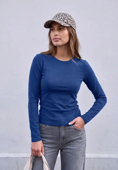 Frau mit blauem Langarmshirt, grauen Jeans und leopardgemusterter Kappe vor neutraler Wand.