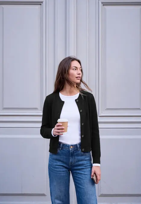 Eine Frau steht vor einer hellen Wand, trägt Jeans, T-Shirt und Cardigan und hält Kaffee sowie Smartphone.