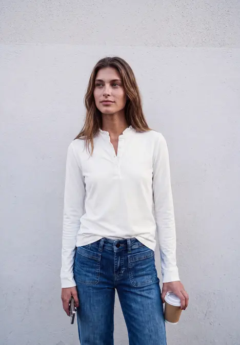 Eine junge Frau in weißem Shirt und Jeans steht vor einer Wand, hält Schlüssel und Kaffee in den Händen.