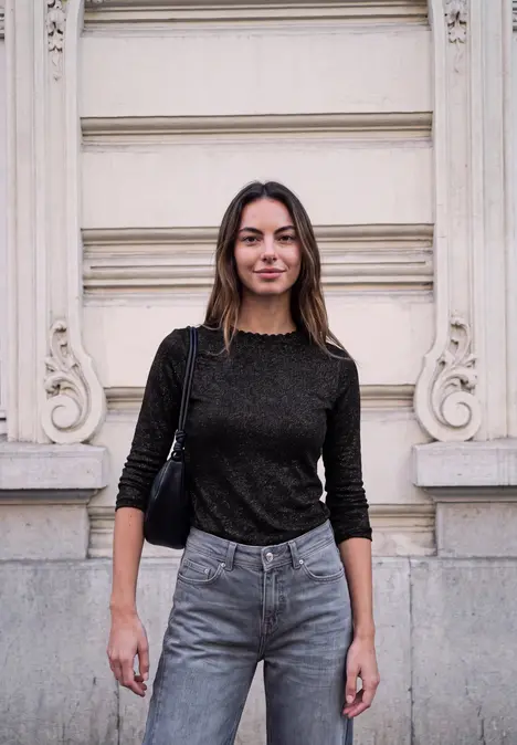Eine Frau in schwarzem Pullover und Jeans steht vor einer hellen Fassade und blickt in die Kamera.
