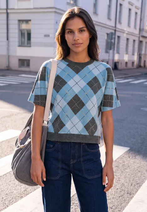 Junge Frau in kariertem Pullover und Jeans steht mit großer Tasche auf sonniger Straßenecke.