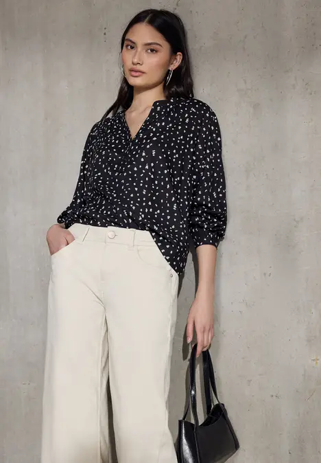 Eine Person trägt eine schwarze Bluse mit weißen Punkten, helle hohe Hosen und hält eine kleine schwarze Tasche.