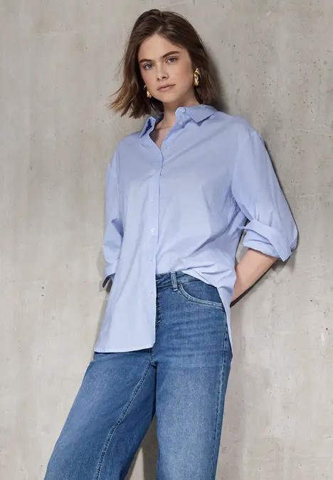 Frau mit lockerem Hemd und Jeans steht vor einer Wand.