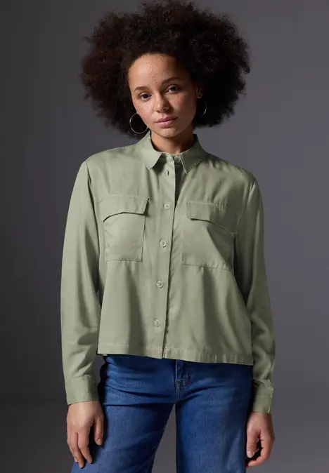 Frau mit Afro trägt grüne Bluse und Jeans, steht vor grauem Hintergrund.