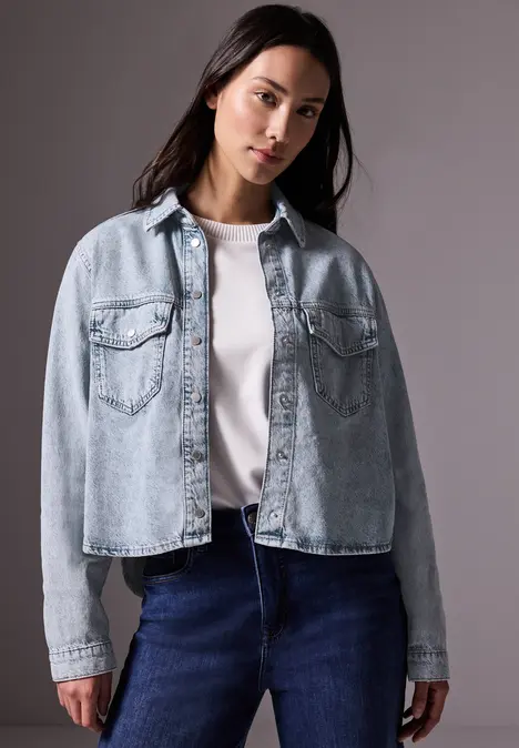 Frau mit Jeansjacke und weißem Shirt steht vor grauem Hintergrund.