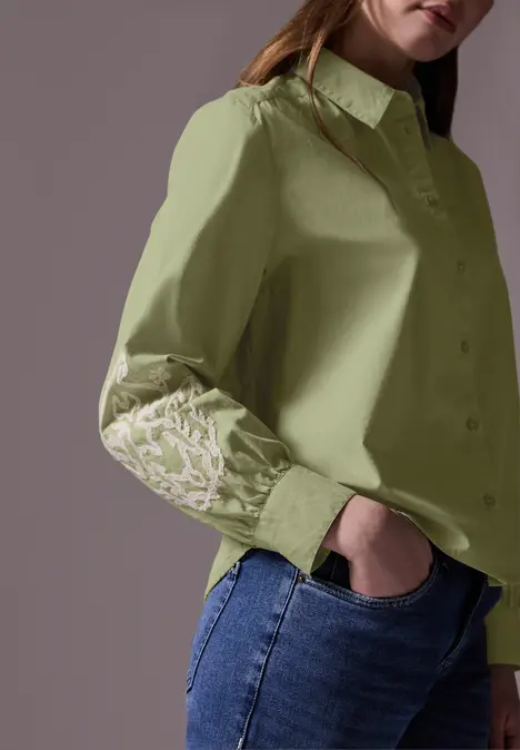 Frau in hellgrüner Bluse mit Stickerei am Ärmel und blauer Jeans.