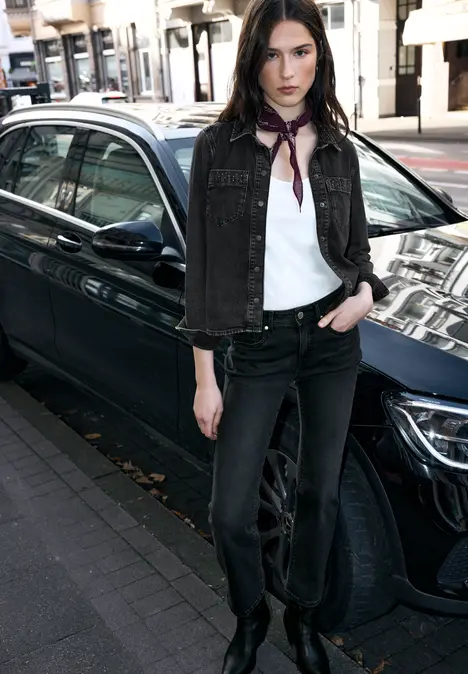 Vrouw in zwarte spijkerjas, wit shirt, zwarte broek en sjaaltje leunend tegen een zwarte auto op straat.