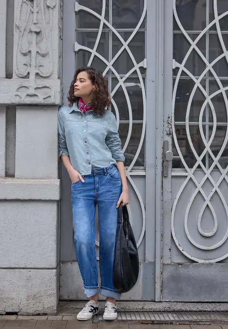 Een vrouw in jeans en blouse poseert nonchalant voor een versierd metalen deur.
