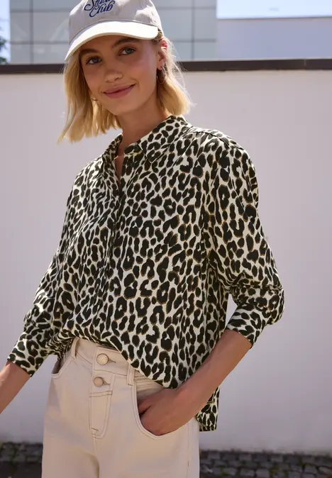Frau mit beige-grüner Leo-Bluse, hellen High-Waist-Hosen und Cap steht vor einer hellen Wand.