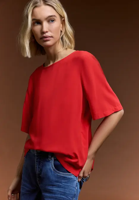 Person steht vor braunem Hintergrund, trägt rotes T-Shirt mit kurzen Ärmeln und blaue Jeans.