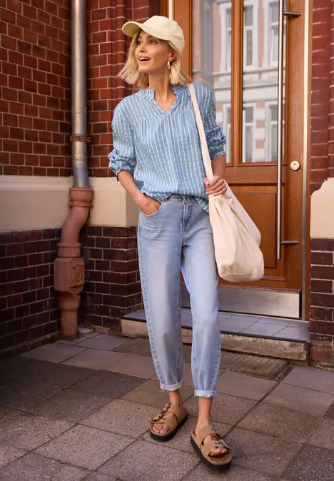 Person vor Hauseingang in blau-weiß gestreiftem Hemd, hellen Jeans, Sandalen und Tragetasche.