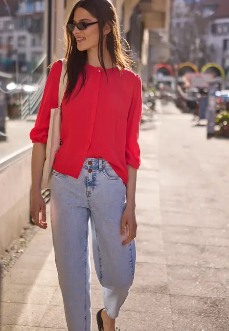 Frau in roter Bluse und blauen Jeans spaziert an sonnigem Tag auf dem Bürgersteig vor Geschäften.