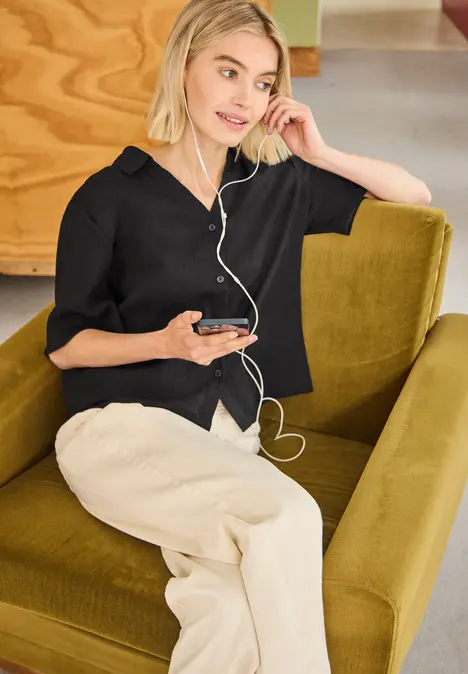 Frau sitzt entspannt im grünen Sessel, hört Musik mit Kopfhörern und hält ein Smartphone.