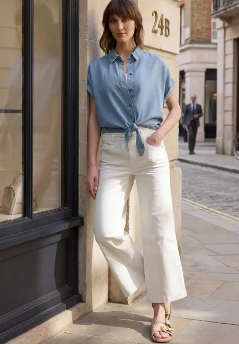 Vrouw in lichtblauw kort overhemd en witte wijde jeans staat op straat.