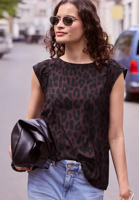Frau mit lockigem Haar, Animal-Print-Shirt, Sonnenbrille und Jeans, hält Lederjacke in der Hand.