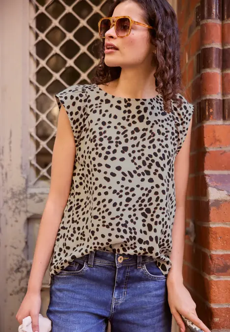 Vrouw leunt nonchalant tegen bakstenen muur, draagt jeans, mouwloos luipaardtop en zonnebril.