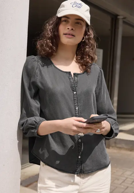 Vrouw in zwarte blouse en witte broek leunt tegen een zuil en houdt een smartphone vast.