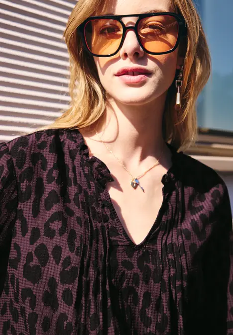 Junge Frau mit Oversize-Sonnenbrille, gemusterter Bluse und zarter Halskette vor Jalousien.
