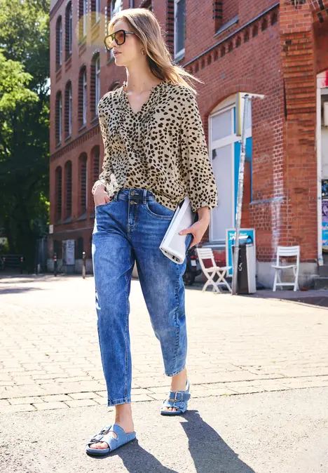 Vrouw in luipaardblouse, wijde jeans en blauwe sandalen wandelt bij zonneschijn langs een bakstenen gebouw.