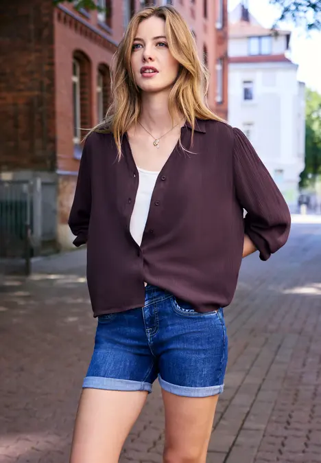 Junge Frau mit weinroter Bluse, weißen Top und Jeans-Shorts steht in einer gepflasterten Straße vor Ziegelgebäuden.