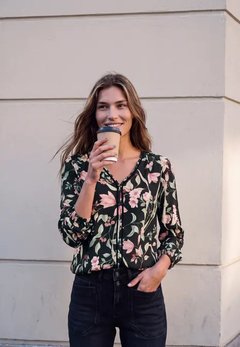 Een glimlachende vrouw in een bloemenblouse en spijkerbroek houdt een koffiebeker voor een muur.