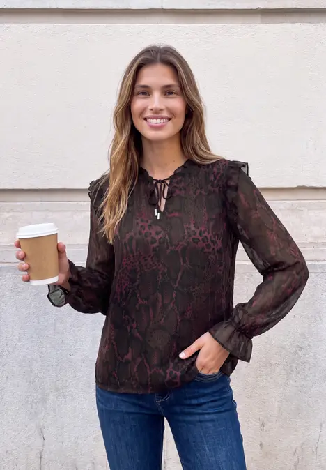 Eine lächelnde Frau in Bluse und Jeans hält einen Pappbecher vor einer hellen Wand.