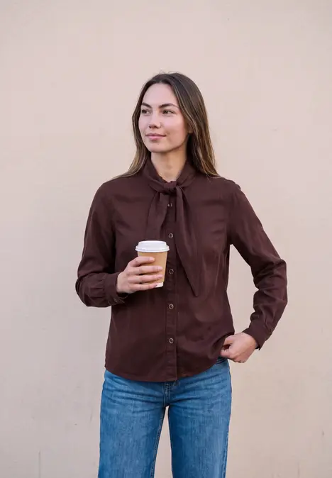 Eine Frau trägt braune Bluse und blaue Jeans, hält einen Kaffee und schaut zur Seite.