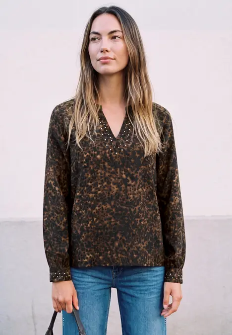 Frau in gemusterter dunkler Bluse und blauen Jeans vor hellem Hintergrund.