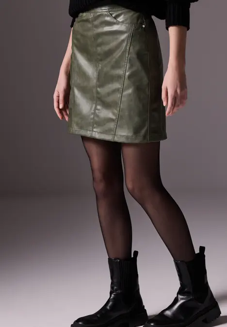 Frau mit grünem Lederrock, schwarzem Pulli, Strumpfhose und Stiefeln.