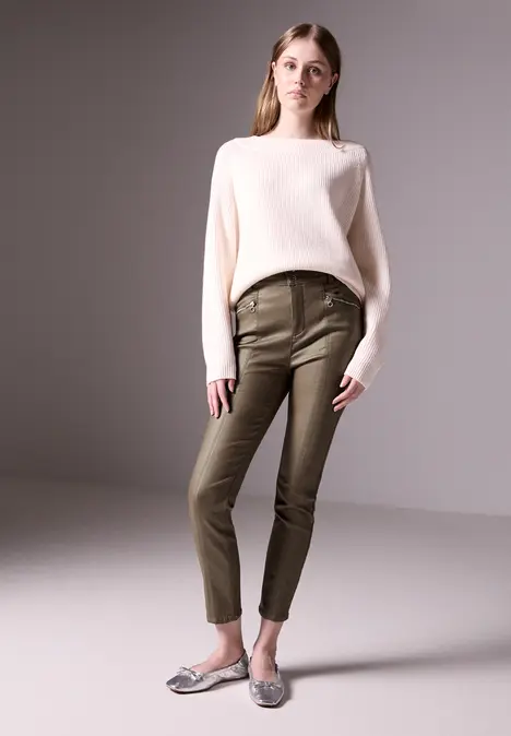 Frau in weißem Pullover, olivgrüner Hose und silbernen Schuhen steht lässig.