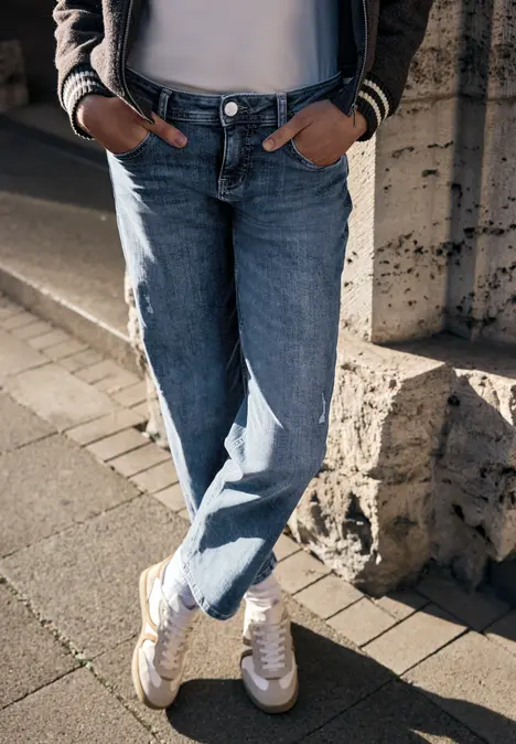 Person in Jeans, weißem Top und Sneakern lehnt an einem Steinpfeiler.