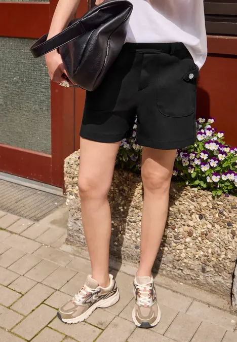 Person steht auf gepflastertem Boden, trägt weiße Bluse, schwarze Shorts, Sneaker und hält eine schwarze Tasche.