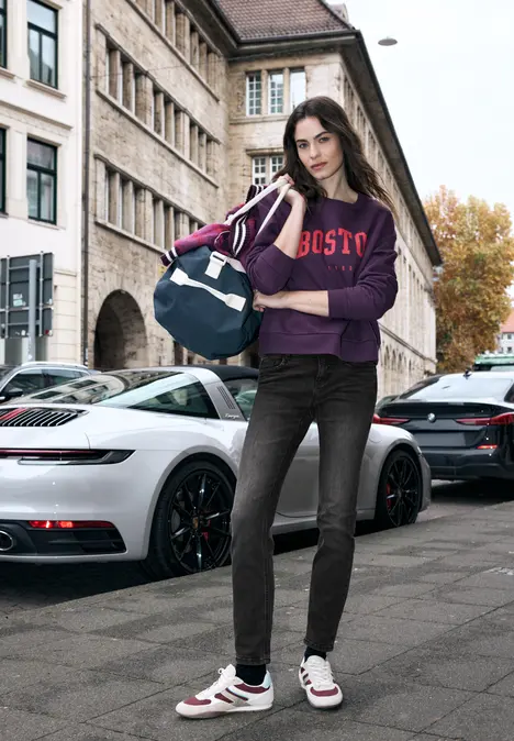 Persoon in paarse hoodie, jeans en sneakers staat met sporttas bij een witte sportwagen op straat.