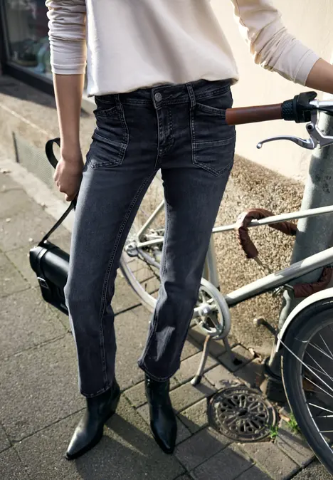 Persoon in zwarte high-waist jeans en zwarte enkellaarsjes, staand naast een fiets op het trottoir.