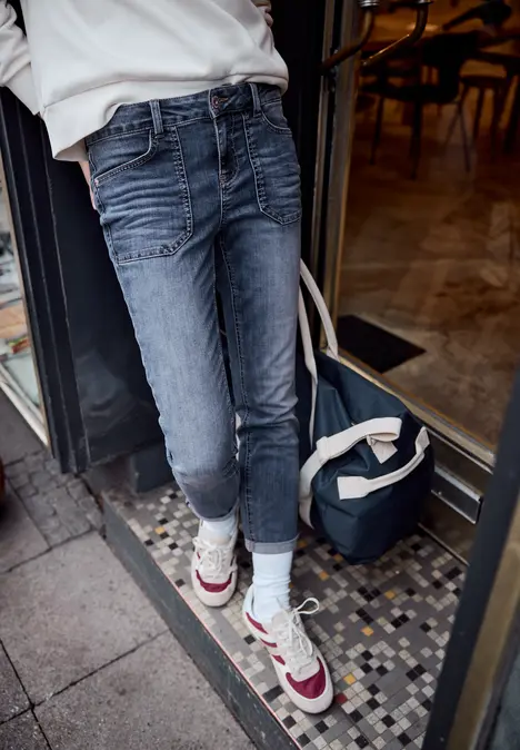 Persoon in blauwe jeans, witte sokken en sneakers met zwarte tas naast de ingang van een café.