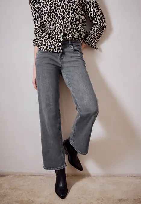 Vrouw in luipaardprint blouse, grijze jeans en zwarte enkellaarzen voor een effen achtergrond.