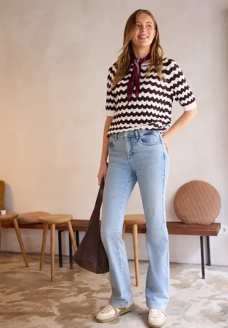 Vrouw in zigzagshirt, wijde jeans en grote tas voor een eenvoudig interieur
