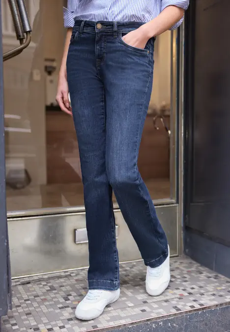 Iemand in donkerblauwe flared jeans en witte sneakers, staand in een deuropening.