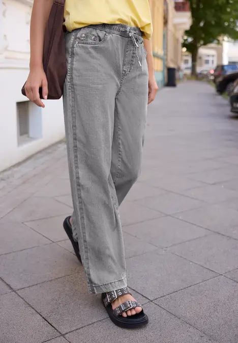 Vrouw in losse grijze jeans en zwarte sandalen loopt op de stoep.