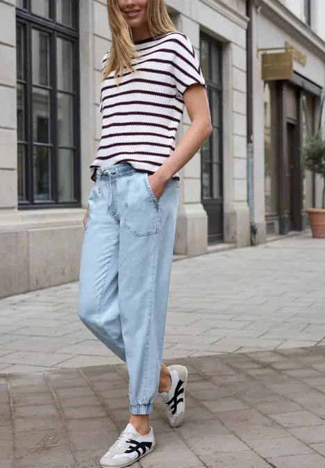Vrouw in gestreept shirt en lichtblauwe jeans, hand in zak, nonchalant lopend over de stadstraat.