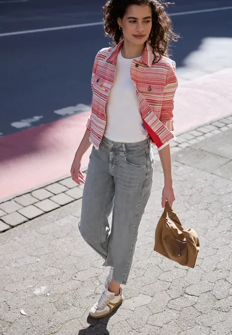 Vrouw in roze gestreepte jas, wit shirt, grijze jeans en gouden schoenen loopt met een bruine tas.