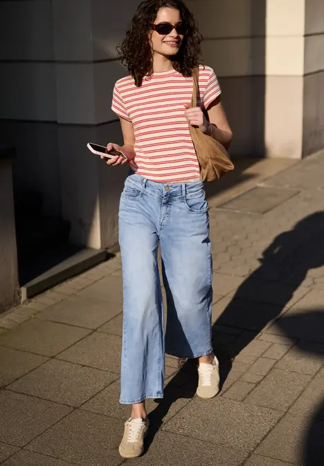 Vrouw in rood-wit gestreept T-shirt, wijde spijkerbroek en sneakers loopt met smartphone over een zonnig trottoir.