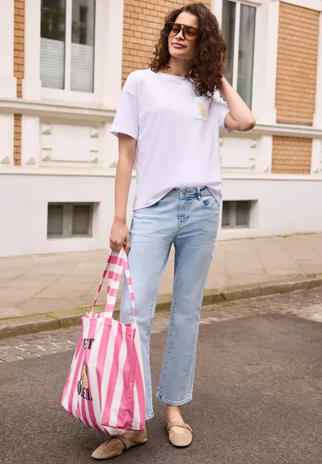 Frau mit lockigem Haar, weißem T-Shirt, Jeans und rosa-weiß gestreifter Tasche steht auf dem Bürgersteig.