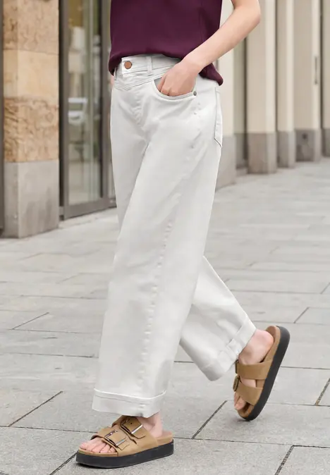 Persoon met witte wijde jeans en bruine sandalen op een geplaveerd trottoir voor een gevel.