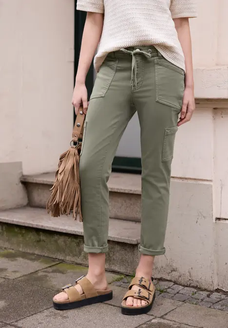 Persoon draagt kaki cargobroek, gestreept shirt, sandalen en franje-tas op bestrating.