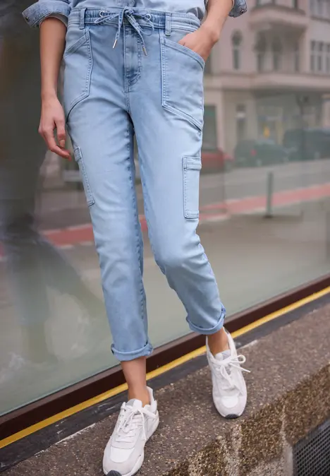 Vrouw in lichte, losse spijkerbroek en witte sneakers staat op de stoep voor een glazen gevel.