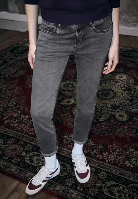 Persoon staat op een gemêleerd tapijt, draagt grijze opgerolde jeans en witte Nike-sneakers met bordeaux accenten.