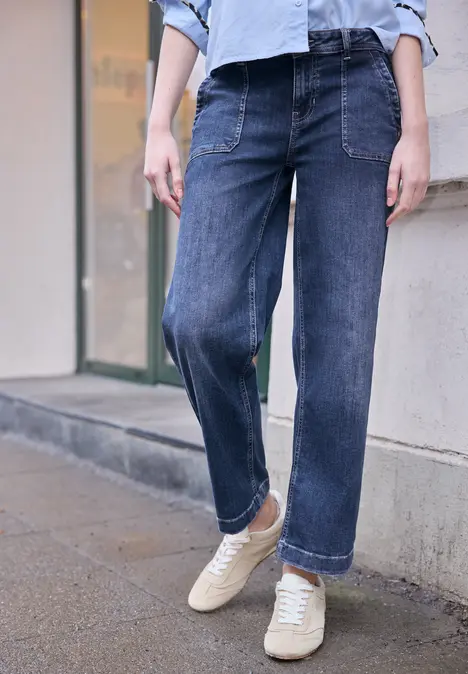 Iemand staat op de stoep voor een gebouw, draagt wijde blauwe jeans en witte sneakers.