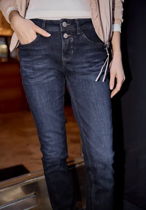 Persoon in donkerblauwe, nauwsluitende jeans en lichte jas, hand in de zak.