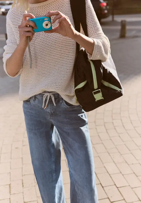 Person in hellem Pullover und Jeans macht Foto mit türkisfarbener Kamera, schwarzer Rucksack an der Schulter.
